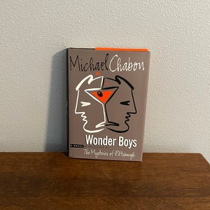 May include: A hardcover book titled "Wonder Boys" by Michael Chabon. The cover is gray with a stylized illustration of two faces and a cocktail glass. The title and author's name are in white and black.