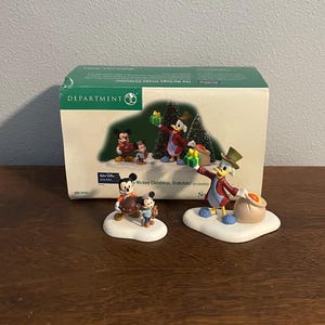 Puede incluir: Un conjunto de dos figuras de cerámica que representan a Mickey Mouse y Donald Duck vestidos con ropa de invierno y sosteniendo regalos de Navidad. Las figuras forman parte de un conjunto "Mickey Christmas, Gratchits!"