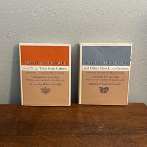 Peut inclure: Deux livres à couverture rigide intitulés "The Juniper Tree and Other Tales from Grimm". Les livres ont une forme rectangulaire avec un motif de blocs de couleur en haut. Un livre a un dessus orange, l'autre un dessus bleu. Le texte sur la couverture comprend le nom de l'auteur.