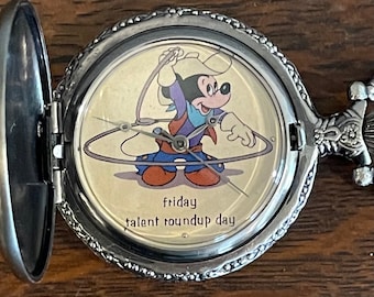 Könnte beinhalten: Eine silberfarbene Taschenuhr mit offenem Deckel, die ein Cartoon-Bild von Mickey Mouse als Cowboy zeigt. Das Zifferblatt der Uhr trägt die Aufschrift "friday talent roundup day". Die Uhr liegt auf einer Holzoberfläche.