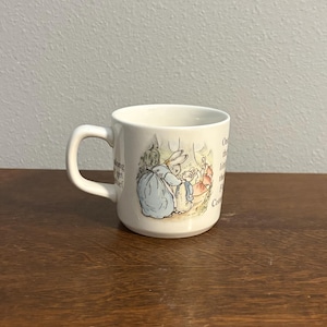 以下が含まれることがあります： 青いドレスを着たウサギと小さなウサギのクラシックなイラストが描かれた白いセラミックマグ。 マグカップには湾曲したハンドルがあり、両側にテキストがあります。 マグカップは木製の表面にあります。