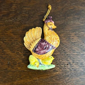 May include: A ceramic hen ornament with a gold string for hanging. The hen is yellow with a maroon and purple accent on its back. Two yellow chicks sit on a green base below the hen.