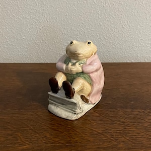 Op de afbeelding: Een keramiek beeldje van een kikker die een roze jas en een groen vest draagt, zittend op een boek. De kikker heeft een lichtbruin lichaam en zwarte ogen.