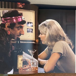 Op de afbeelding: Een barsituatie met twee mensen die tegenover elkaar zitten. E&eacute;n persoon draagt een hoofdband met pailletten en een donkerpaarse outfit. Een fles drank en een cocktailglas staan op de bar. De andere persoon draagt een beige top.