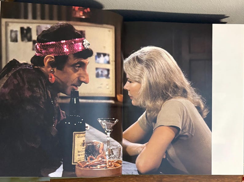 Op de afbeelding: Een barsituatie met twee mensen die tegenover elkaar zitten. E&eacute;n persoon draagt een hoofdband met pailletten en een donkerpaarse outfit. Een fles drank en een cocktailglas staan op de bar. De andere persoon draagt een beige top.