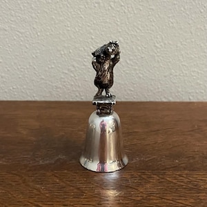 May include: A silver bell with a small bear figurine on top. The bear is holding a small bag in its paws.