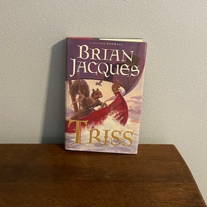 Könnte beinhalten: Ein Hardcover-Buch von Brian Jacques mit dem Titel "Triss". Das Cover zeigt ein Eichhörnchen in einem roten Boot unter einem lila Himmel mit einem weißen Segel. Das Buch liegt auf einer Holzoberfläche vor einer grauen Wand.