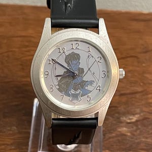 May include: A silver wristwatch with a black leather band. The watch face features a white background with black Roman numeral hour markers and a cartoon image of a boy with a wand. The watch face also includes the numbers 1 through 12.