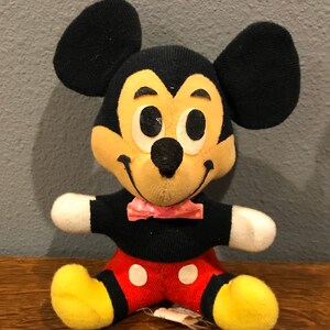 May include: A vintage Mickey Mouse soft toy. The plush has a black body, large black ears, a yellow face, and red shorts with white buttons. It has a pink bow tie and yellow feet. The toy is sitting on a wooden surface.