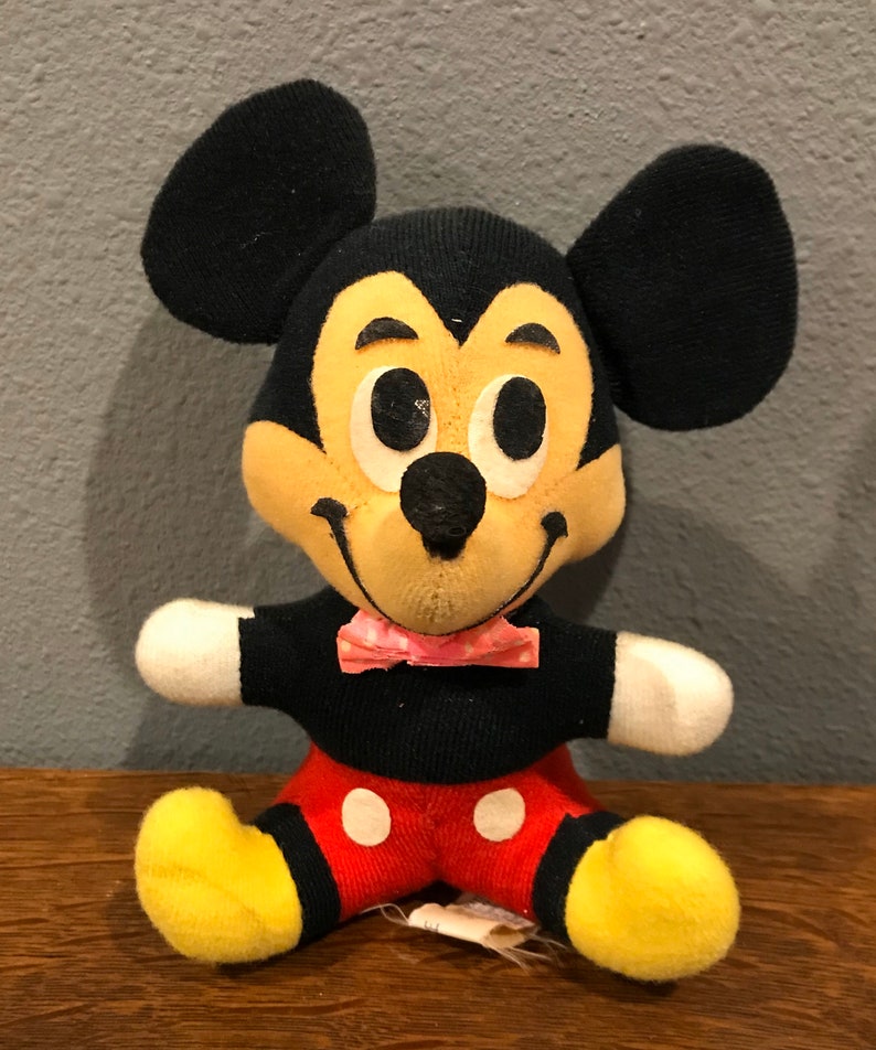 May include: A vintage Mickey Mouse soft toy. The plush has a black body, large black ears, a yellow face, and red shorts with white buttons. It has a pink bow tie and yellow feet. The toy is sitting on a wooden surface.