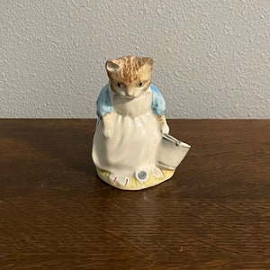 Może przedstawiać: Ceramiczna figurka kota w niebieskiej sukience, trzymającego wiadro. Kot ma brązowe futro i biały fartuch. Figurka stoi na żółtej podstawie z zieloną trawą.