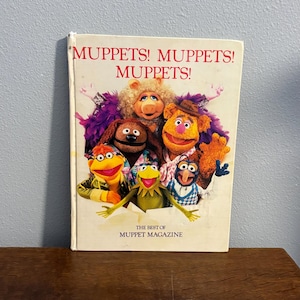 May include: A vintage copy of "Muppets! Muppets! Muppets!" magazine. The cover features a colourful group of Muppet characters, including Kermit the Frog, Miss Piggy, and Fozzie Bear. The magazine is on a wooden surface.