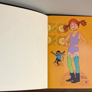 May include: A paper doll book page featuring a Pippi Longstocking character with red pigtails, wearing a purple leotard, one green and one blue striped stocking, and black boots. A small monkey and a floral pattern are printed on the orange background.