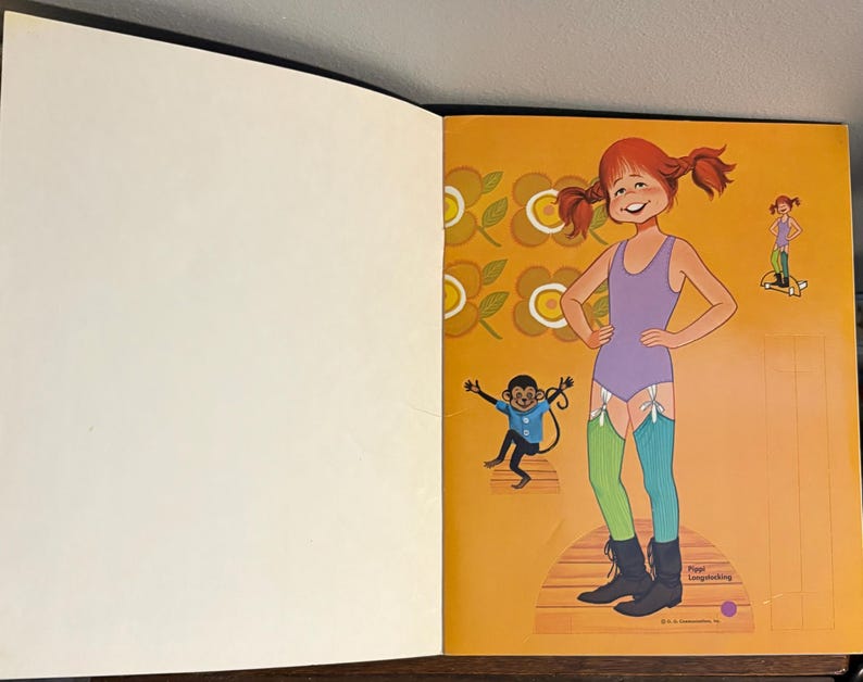 May include: A paper doll book page featuring a Pippi Longstocking character with red pigtails, wearing a purple leotard, one green and one blue striped stocking, and black boots. A small monkey and a floral pattern are printed on the orange background.