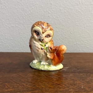 Puede incluir: Figura de cerámica de un búho y una ardilla. El búho tiene plumas marrones y crema y ojos grandes. La ardilla es de color marrón rojizo. Ambos están posados sobre una base verde con hierba.