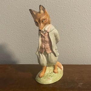 Puede incluir: Figura de cerámica de un zorro vestido con una chaqueta gris claro, un chaleco rosa y pantalones verde claro. El zorro está de pie sobre una base de hierba verde. El zorro tiene pelaje marrón y canela.