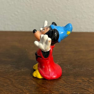 May include: A small figurine of Mickey Mouse in a sorcerer's outfit. The figure has a red robe, a blue hat with a yellow star, and white gloves. The figure is posed with its arms raised.