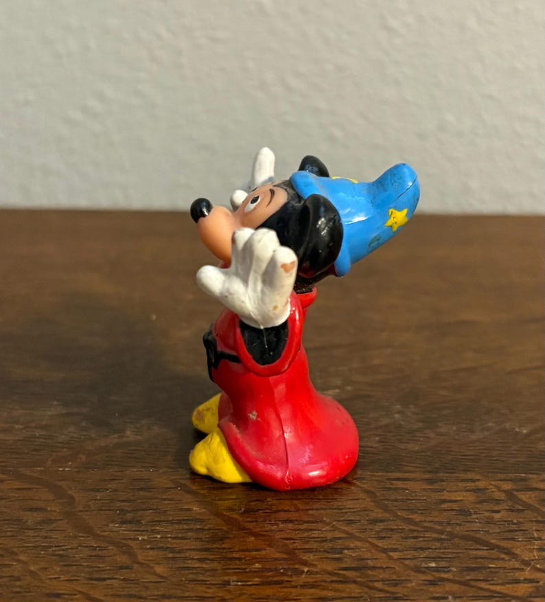 May include: A small figurine of Mickey Mouse in a sorcerer's outfit. The figure has a red robe, a blue hat with a yellow star, and white gloves. The figure is posed with its arms raised.