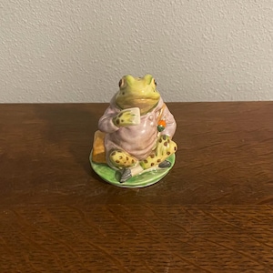 Op de afbeelding: Een keramieken kikkerfiguur, zittend en een kopje vasthoudend, draagt een roze jas met gele stippenbroek. De kikker staat op een groene basis met een kleine oranje wortel. De figuur heeft een grillige, sprookjesachtige esthetiek.