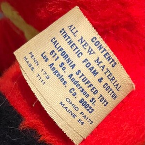 May include: Close-up of a tag with text that reads "ALL NEW MATERIAL SYNTHETIC FOAM & COTTON CALIFORNIA STUFFED TOYS". The tag is beige with blue text and is set against a red and black background.