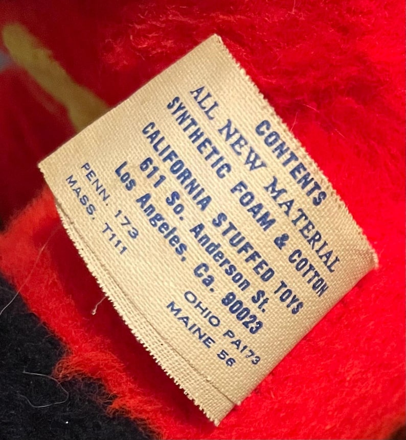 May include: Close-up of a tag with text that reads "ALL NEW MATERIAL SYNTHETIC FOAM & COTTON CALIFORNIA STUFFED TOYS". The tag is beige with blue text and is set against a red and black background.