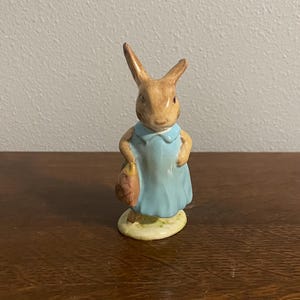 May include: A small ceramic figurine of a brown rabbit wearing a blue dress and holding a brown basket. The rabbit is standing on a green base.