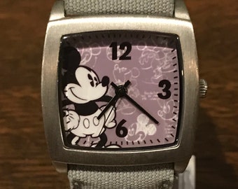 May include: A silver watch with a square face and a gray canvas band. The face has a purple background with a black and white image of Mickey Mouse and a pattern of Mickey Mouse heads. The watch face has black numbers 12, 3, and 6.