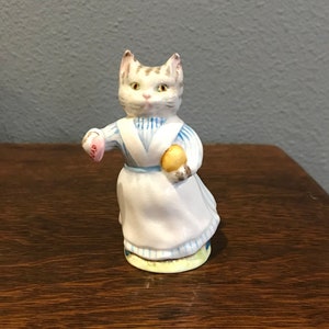 Puede incluir: Figura de cerámica de un gato vestido con un vestido a rayas azules y blancas y un delantal blanco. El gato sostiene un jabón rosa y un objeto amarillo. La figura tiene un diseño pintado detallado y está sobre una base verde.