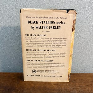 May include: Vintage paperback book from the Black Stallion series by Walter Farley. The cover displays text including the titles of the first three books in the series. The book is on a wooden surface.