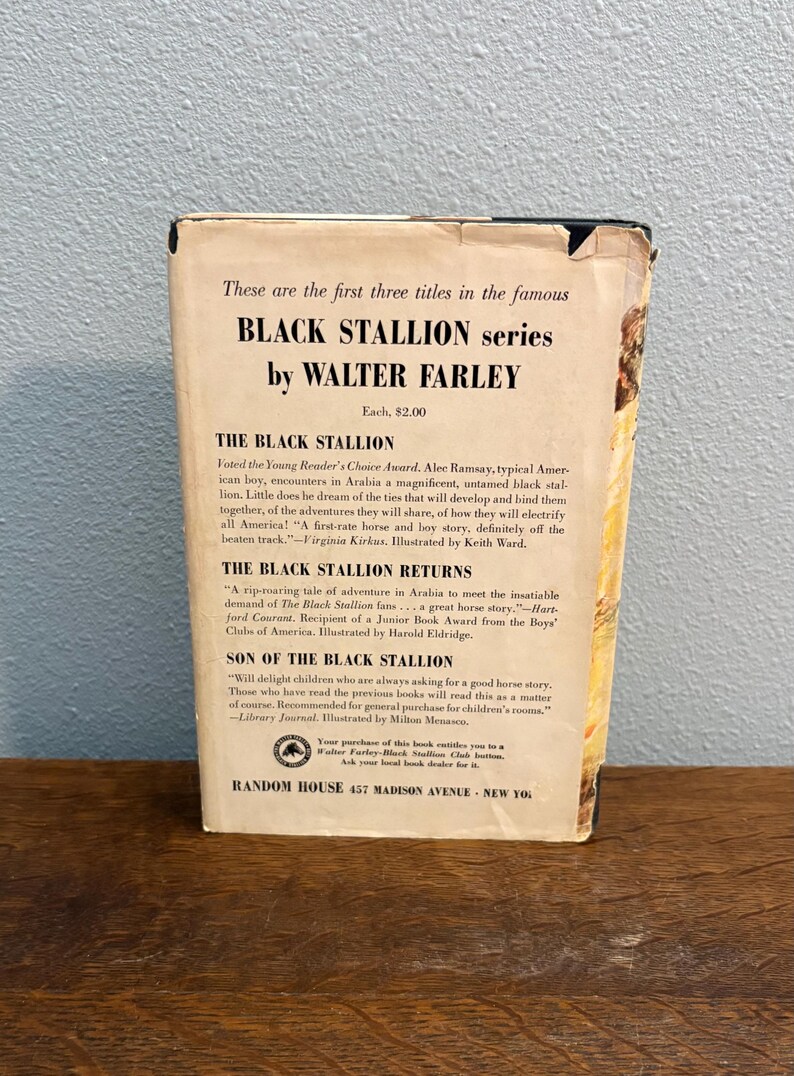 May include: Vintage paperback book from the Black Stallion series by Walter Farley. The cover displays text including the titles of the first three books in the series. The book is on a wooden surface.