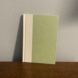 May include: A hardback book with a pale green cover and a cream spine. The cover has an embossed design. The book is on a wooden surface.