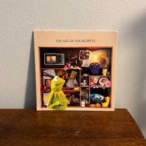 May include: A book cover titled "The Art of the Muppets" showing a shelf display of Muppet characters and props. Kermit the Frog is in the foreground. Other characters include Cookie Monster and a purple monster.