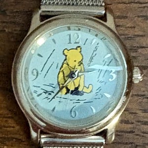 May include: A gold-toned wristwatch with a light blue face featuring a yellow Winnie the Pooh character. The watch has a silver mesh band and displays the numbers 12, 3, 6, and 9.