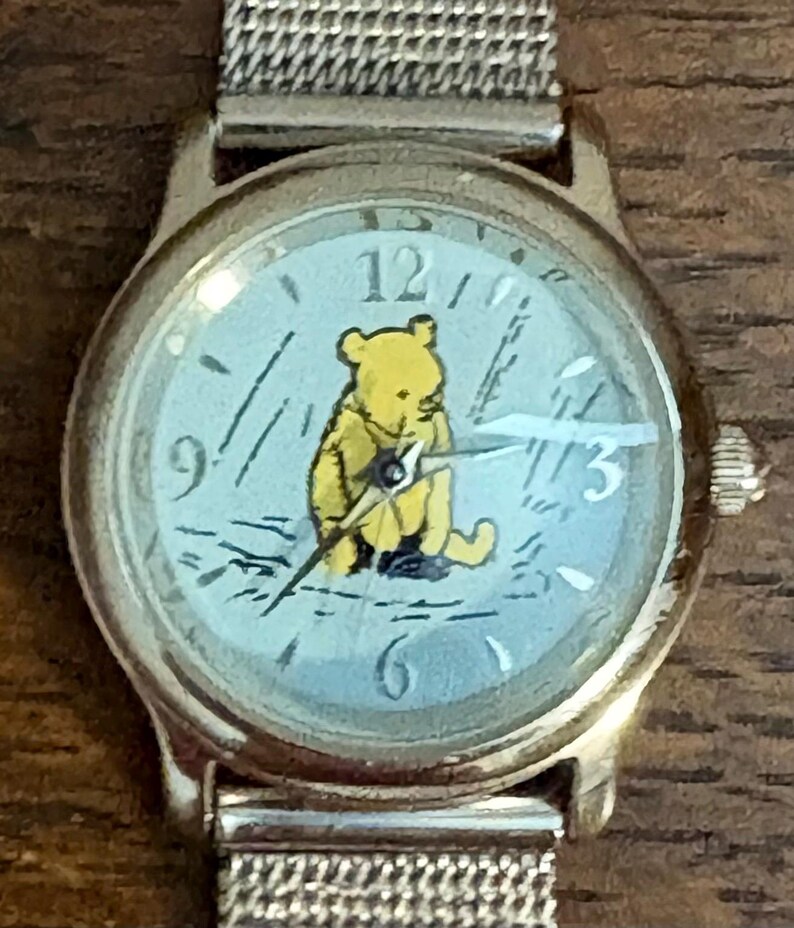 May include: A gold-toned wristwatch with a light blue face featuring a yellow Winnie the Pooh character. The watch has a silver mesh band and displays the numbers 12, 3, 6, and 9.