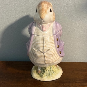 Op de afbeelding: Een keramieken konijnfiguur met een paarse jas en vest. Het konijn heeft een bruin gezicht en staat op een groene basis. De jas heeft gouden knopen en het vest heeft een gestreept patroon.