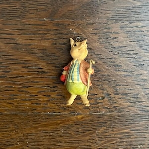 May include: A small, hand-painted figurine of a pig wearing a red, blue, and white striped shirt and green pants. The pig is holding a stick in its right hand and has a red apple in its left hand.