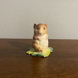 May include: A ceramic figurine of a standing squirrel with a light brown body and a cream-colored belly. The squirrel is perched on a green and yellow leaf base. The figurine is set on a dark brown wooden surface.