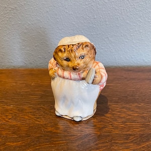May include: A ceramic figurine of a hedgehog wearing a white apron and a red and white checkered shirt. The hedgehog is holding a small, round object in its right hand.