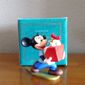 May include: A ceramic figurine of Mickey Mouse carrying a red gift box with a green bow. The figurine is on a green base and is part of a Disney Classics collection.