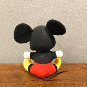 May include: A plush Mickey Mouse doll, viewed from the back, with black ears and head, a black torso, red shorts, yellow feet, and a black tail. The doll is sitting on a wooden surface against a grey wall.