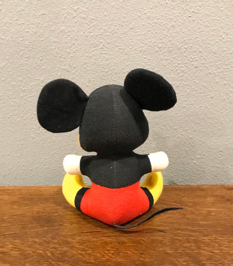 May include: A plush Mickey Mouse doll, viewed from the back, with black ears and head, a black torso, red shorts, yellow feet, and a black tail. The doll is sitting on a wooden surface against a grey wall.