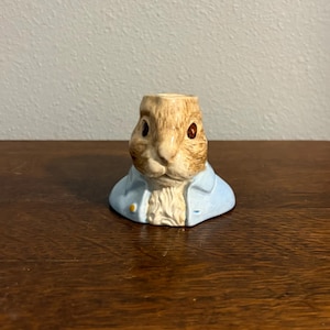 Może przedstawiać: Ceramiczna figurka królika w jasnoniebieskiej kurtce. Królik ma brązowe futro, brązowe oczy i białą pierś. Figurka jest przedmiotem dekoracyjnym, być może świecznikiem lub małym wazonem.