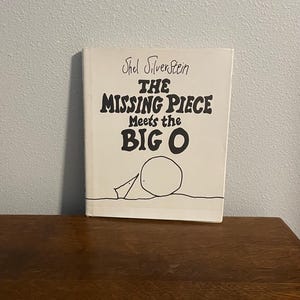 May include: A white book with black text that reads "Shel Silverstein The Missing Piece Meets the Big O". The cover features a simple black and white illustration of a circle with a smaller shape next to it.