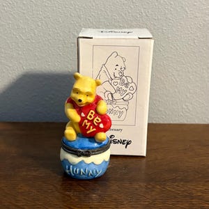 May include: A ceramic Winnie the Pooh figurine sitting on a blue and white honey pot base. The yellow bear holds a red heart with the text Be My. The figurine is displayed in front of its original white cardboard box featuring a line drawing.