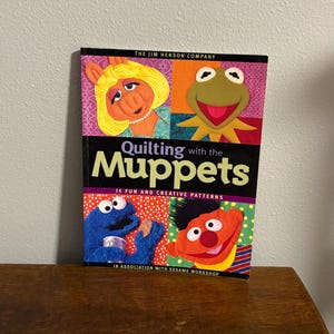 May include: A book titled "Quilting with the Muppets" featuring images of Miss Piggy, Kermit the Frog, Cookie Monster, and Ernie. The cover includes the text "15 Fun and Creative Patterns" and "In Association with Sesame Workshop."