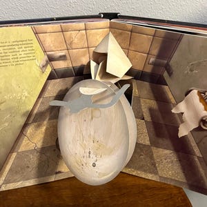 May include: Open book with a pop-up illustration of a bathroom scene. The pop-up features a large, oval-shaped object with a gray silhouette. The book's pages display text and a tiled floor design.