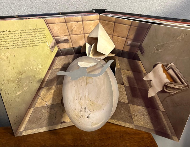 May include: Open book with a pop-up illustration of a bathroom scene. The pop-up features a large, oval-shaped object with a gray silhouette. The book's pages display text and a tiled floor design.