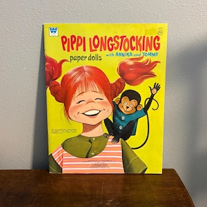 May include: A vintage Pippi Longstocking paper dolls book with a bright yellow cover. The illustration features a red-haired character with pigtails and a small monkey on their shoulder. The text reads Pippi Longstocking paper dolls with Annika and Tommy.