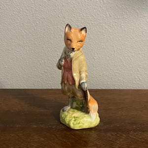 May include: A ceramic fox figurine wearing a tan coat, burgundy waistcoat, and brown trousers, holding a walking stick. The fox stands on a green and yellow base. The fox has orange fur and black detailing.