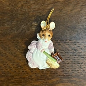 May include: A ceramic mouse ornament, dressed in a pink and white outfit, holding a broom and a small brown bucket. The mouse has brown fur and large ears. A gold string is attached for hanging. The figurine is likely a Christmas decoration.